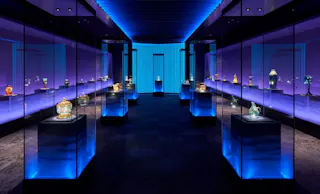 A dimly lit museum gallery displays ornate glass and ceramic artifacts in glass cases, illuminated by blue and purple lighting with a glowing wall at the far end.