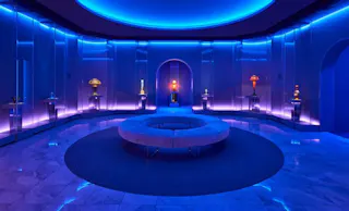 A modern, circular room with blue lighting features a central round bench, marble floors, mirrored walls, and art pieces displayed on pedestals around the perimeter. An arched doorway is visible in the back.