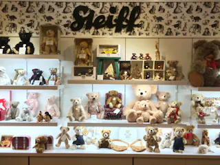 A display of various Steiff stuffed animals, mainly teddy bears, arranged on shelves with a patterned backdrop and a large “Steiff” sign above. The shelves also include other plush animals and small figurines.