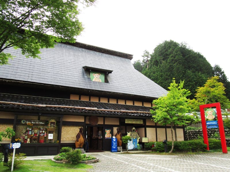 Traditional Japanese building with a dark tiled roof, surrounded by green trees. There are bear cartoon decorations on the building and sign, and a red torii gate on the right. The setting appears peaceful and inviting.