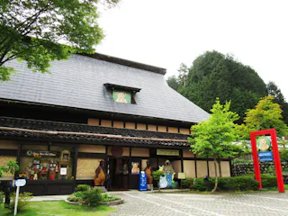 Traditional Japanese building with a dark tiled roof, surrounded by green trees. There are bear cartoon decorations on the building and sign, and a red torii gate on the right. The setting appears peaceful and inviting.