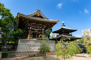 A traditional Japanese temple with ornate wooden architecture stands beside a bell tower, surrounded by trees and greenery under a clear blue sky. Modern buildings are visible in the background.