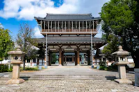 A traditional Japanese temple with a large wooden gate, intricate roof details, stone lanterns on either side, and trees surrounding the entrance on a sunny day.