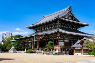 A large traditional Japanese temple with ornate wooden architecture and a gray-tiled roof stands under a clear blue sky, surrounded by a gravel courtyard and some greenery.