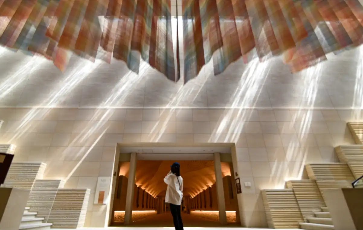 A person stands in a large, modern hall with light streaming through colorful, hanging fabric panels, casting patterns on the walls and floor. The space features geometric steps and a well-lit archway in the background.