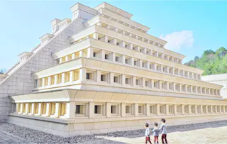 Two young children wearing face masks stand in front of a large, modern stepped pyramid structure with rectangular windows, under a bright blue sky with some greenery in the background.