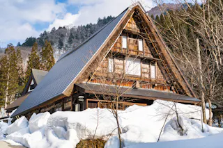 A traditional wooden house with a steep thatched roof stands surrounded by deep snow and bare trees, with forested mountains in the background under a partly cloudy sky.