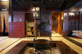 A traditional Japanese room with tatami mats, a kettle hanging over a sunken hearth, and a fish-shaped metal ornament above it. In the background are wooden furniture, shelves with books, and sliding doors.