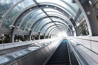 A modern, covered outdoor escalator with a curved glass and metal roof ascends toward bright natural light, surrounded by trees visible through the transparent panels.