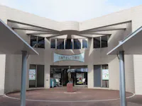 The entrance of a modern building with tiled walls, large glass doors, and a sculpture of a person in front. Signs with cartoon faces and Japanese text are displayed next to the doors.