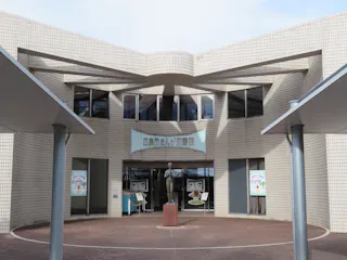The entrance of a modern building with tiled walls, large glass doors, and a sculpture of a person in front. Signs with cartoon faces and Japanese text are displayed next to the doors.