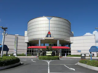A modern building with a large cylindrical section above the entrance, glass windows, a red awning, and manicured bushes in front. The sky is clear and blue.