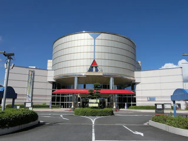 A modern building with a large cylindrical section above the entrance, glass windows, a red awning, and manicured bushes in front. The sky is clear and blue.
