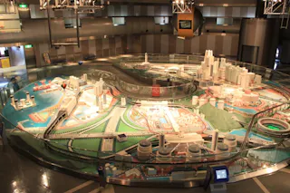 A large, detailed scale model of an urban city is displayed indoors, featuring miniature buildings, roads, bridges, and waterways, all enclosed in a glass case under bright lighting.