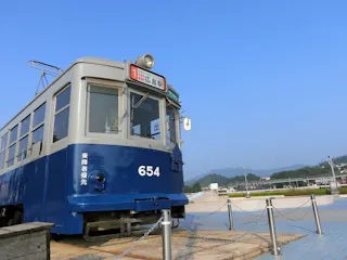 A vintage blue and gray streetcar numbered 654 is displayed outdoors on a sunny day, surrounded by chains and posts, with mountains and greenery visible in the background.