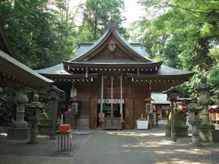 A traditional Japanese Shinto shrine with a wooden structure, stone lanterns, and trees surrounding the building. Decorative ropes and paper streamers hang at the entrance.
