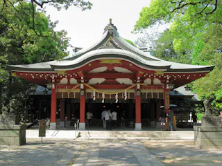 A traditional Japanese Shinto shrine with a curved roof, red pillars, and decorative elements. People are entering under the shaded entrance, surrounded by green trees and stone lanterns.