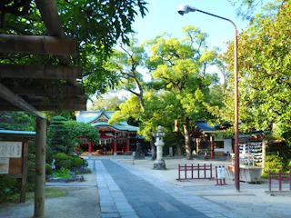 A stone path leads to a traditional Japanese shrine surrounded by lush green trees, lanterns, and manicured gardens, with a red and white building visible in the background under bright daylight.