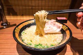 A bowl of ramen with creamy broth, noodles, sliced pork, and green onions sits on a wooden table. Chopsticks lift some noodles from the bowl, ready to eat.