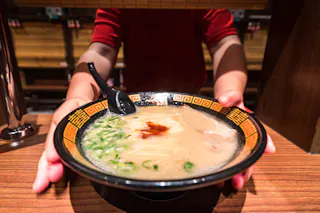 A person in a red shirt serves a bowl of ramen with green onions and a dollop of chili paste in a patterned bowl, holding it above a wooden counter with a black spoon inside.