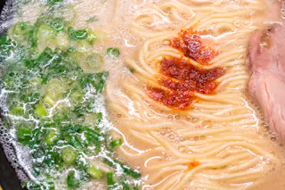 Close-up of ramen noodles in broth with chopped green onions, a dollop of red chili paste, and a slice of meat, seen bubbling in a hot soup.