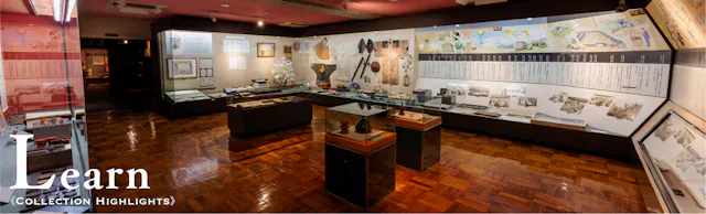 A museum exhibit room with glass display cases holding artifacts, documents, and photos. Informational panels and historical items are showcased on walls and in cabinets. The word "Learn" appears in large text at the bottom left.