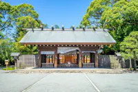 A traditional Japanese Shinto shrine with a wooden gate, surrounded by a fence and lush green trees under a clear blue sky.