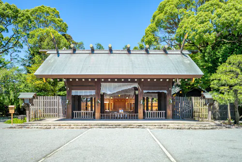 A traditional Japanese Shinto shrine with a wooden gate, surrounded by a fence and lush green trees under a clear blue sky.
