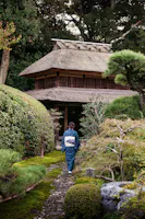 A person wearing a traditional kimono walks along a stone path through a lush Japanese garden towards a wooden gate with a thatched roof, surrounded by greenery and trees.