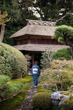 A person wearing a traditional kimono walks along a stone path through a lush Japanese garden towards a wooden gate with a thatched roof, surrounded by greenery and trees.