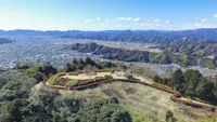 Aerial view of a scenic hillside park with manicured lawns, paths, and a small pavilion, overlooking a sprawling city and surrounded by mountains under a clear blue sky.