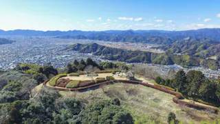 Aerial view of a scenic hillside park with manicured lawns, paths, and a small pavilion, overlooking a sprawling city and surrounded by mountains under a clear blue sky.