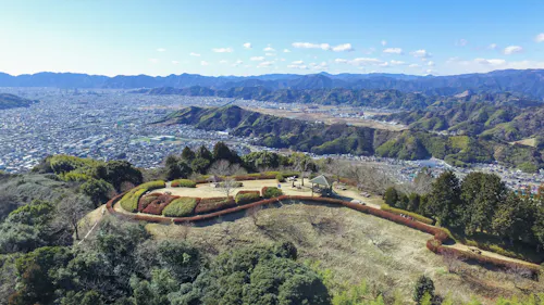 Aerial view of a scenic hillside park with manicured lawns, paths, and a small pavilion, overlooking a sprawling city and surrounded by mountains under a clear blue sky.