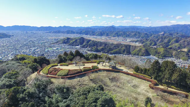 Aerial view of a scenic hillside park with manicured lawns, paths, and a small pavilion, overlooking a sprawling city and surrounded by mountains under a clear blue sky.