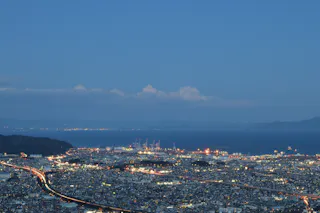 A cityscape at dusk with a brightly lit port near the coastline, roads illuminated by car lights, and calm blue water with mountains faintly visible in the background under a clear sky.