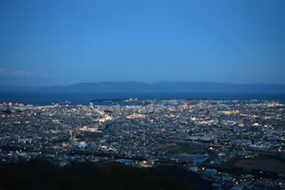 Aerial view of a city at dusk with buildings illuminated by artificial lights, surrounded by water and distant mountains under a clear blue sky.