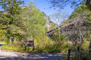 A rustic building surrounded by dense green trees and shrubs under a clear blue sky, with a wooden signpost featuring Japanese text near a gravel path in a mountainous area.