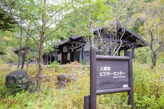 A wooden sign in Japanese stands in front of the Kamikochi Visitor Center, a modern building surrounded by lush green trees and vegetation in a natural setting.