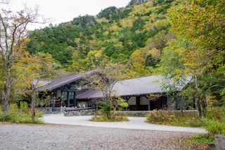 A modern building with sloped roofs sits among trees with early autumn foliage, set against a lush, green hillside. A gravel path leads to the entrance, and the surroundings are calm and natural.