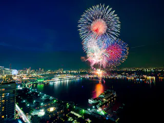 Colorful fireworks explode in the night sky above a city waterfront, reflecting on the water below. City lights, buildings, and a docked ship are visible in the illuminated urban landscape.