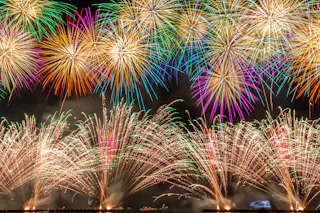 Colorful fireworks explode in the night sky, creating vibrant patterns of red, green, yellow, purple, and white above a dark landscape, illuminating the scene with bright, festive light.