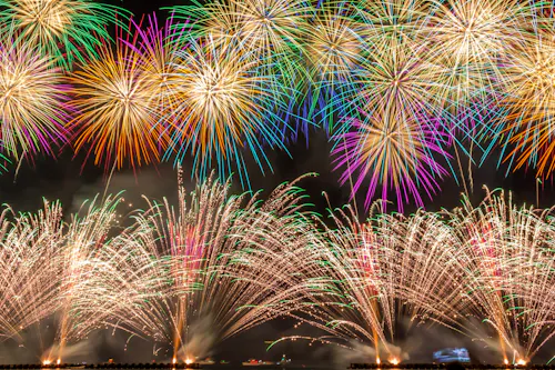Colorful fireworks explode in the night sky, creating vibrant patterns of red, green, yellow, purple, and white above a dark landscape, illuminating the scene with bright, festive light.