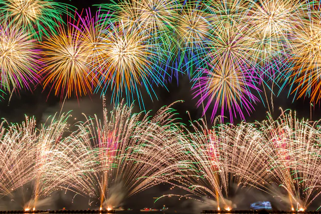 Colorful fireworks explode in the night sky, creating vibrant patterns of red, green, yellow, purple, and white above a dark landscape, illuminating the scene with bright, festive light.