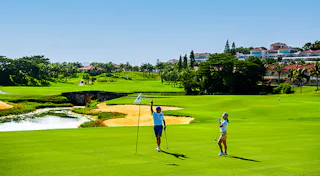 Two people stand on a lush green golf course near a flag and hole, with a small bridge, pond, and resort buildings in the background under a clear blue sky.