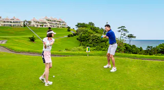 Two people on a green golf course, preparing to swing their clubs. The ocean and trees are in the background, with resort buildings visible on the hillside. The weather is sunny and clear.