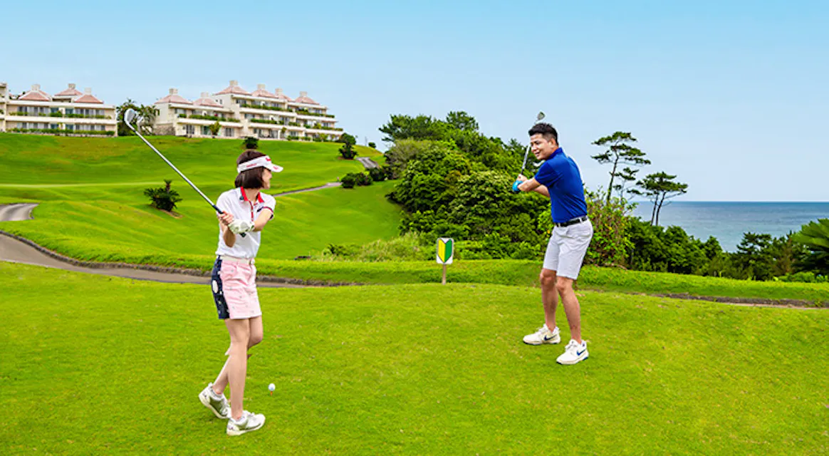 Two golfers teeing off on a sunny fairway at Kanucha Golf Course in Okinawa, with resort buildings and the sea in the background