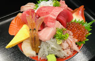 A colorful bowl of assorted sashimi including tuna, salmon, shrimp, tamago, ikura (salmon roe), raw fish, shrimp, and garnishes, presented on rice with a wasabi dollop.