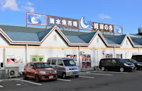 A row of cars is parked in front of a white building with teal roofs and Japanese signs. The signs feature fish illustrations and blue text. The sky is clear with a few clouds.