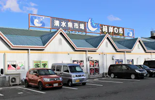 A row of cars is parked in front of a white building with teal roofs and Japanese signs. The signs feature fish illustrations and blue text. The sky is clear with a few clouds.