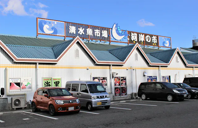 A row of cars is parked in front of a white building with teal roofs and Japanese signs. The signs feature fish illustrations and blue text. The sky is clear with a few clouds.